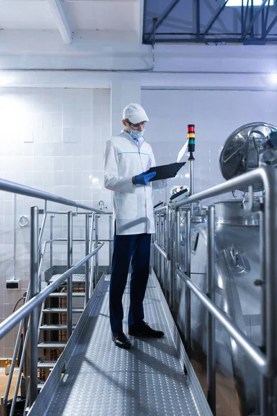 Inspektor w masce i peeling stoi z folderem-tablet w dłoniach w zakładzie mleczarskim — Zdjęcie stockowe