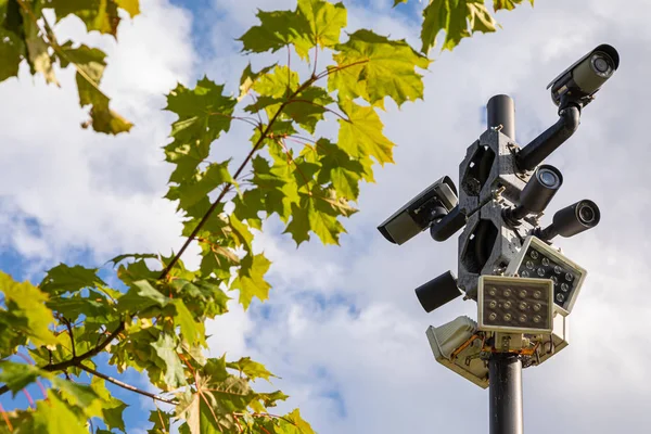 Câmeras de vigilância pretas e lâmpadas de rua brancas em uma coluna contra o céu e folhas verdes de árvore de bordo — Fotografia de Stock