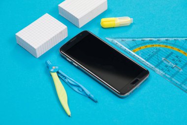 İki beyaz çizik kağıdı, bir çift pusula, bir cetvel, sarı metin işareti ve mavi arka planda siyah akıllı telefon yalıtılmış