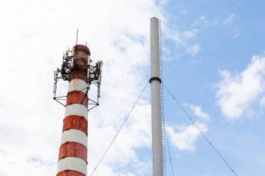 Üstünde çelik döşemesi olan kırmızı beyaz baca ve yanında da mavi gökyüzünde kürklü bulutlar olan ağaç.