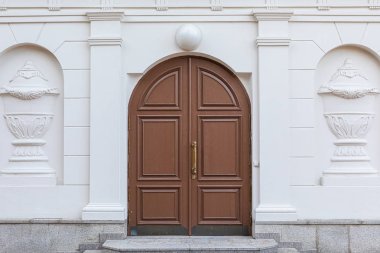 Çift kahverengi, ahşap kemerli, üzerinde lamba olan, beyaz duvarlı, kabartmalı ve granit kaplama basamaklı bir kapı.