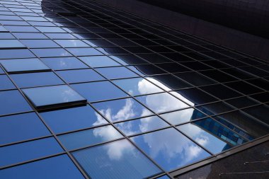 Binanın mavi cam yüzeyinin geniş açılı görüntüsü mavi gökyüzünü beyaz bulutlarla yansıtıyor. Çok az pencere açılır