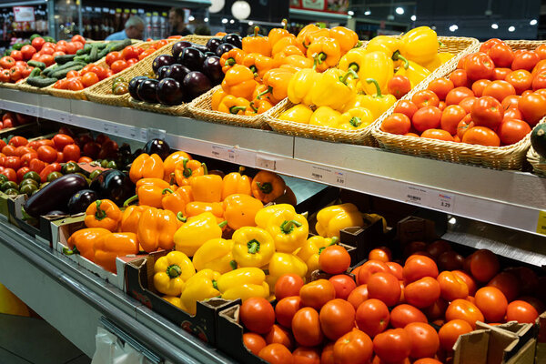 Angular view of supermarket counter with wicker baskets and lug boxes with fresh tomatoes, orange and yellow peppers and eggplants