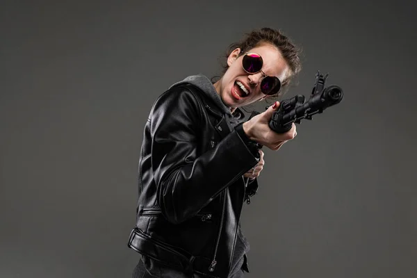 fashionable young woman in leather jacket and sunglasses posing with assault rifle against dark background