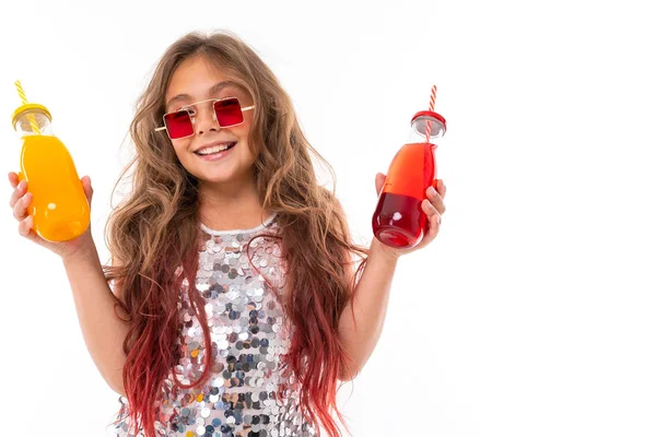 Smiling girl in sparkling dress and stylish square red sunglasses, holding two glass bottles: with red drink and with yellow drink isolated