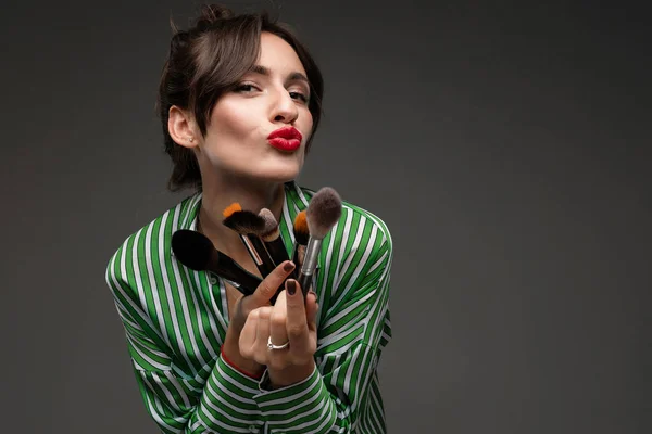 Portrait of young woman making duck face wearing blue and white striped blouse holding makeup brushes in her hands isolated. Photo with depth of field