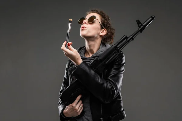 fashionable young woman in leather jacket and sunglasses posing with assault rifle against dark background
