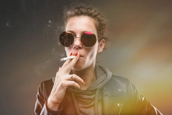 fashionable young woman in leather jacket and sunglasses posing with cigarette against dark background