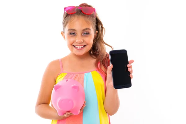 在白色背景下摆出可爱的小女孩的姿势 拿着小猪银行和手机 — 图库照片