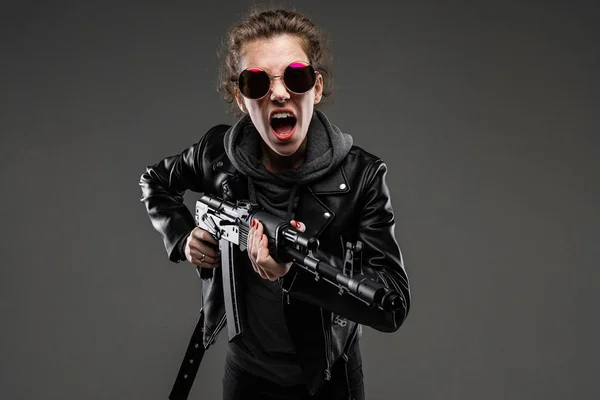 fashionable young woman in leather jacket and sunglasses posing with assault rifle against dark background