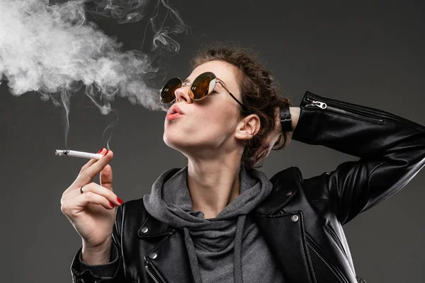 fashionable young woman in leather jacket and sunglasses posing with cigarette against dark background