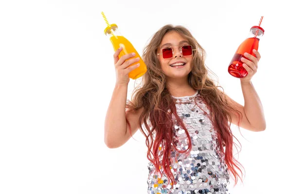 Happy girl in sparkling dress and stylish square red sunglasses, holding two glass bottles: with red drink and with yellow drink isolated