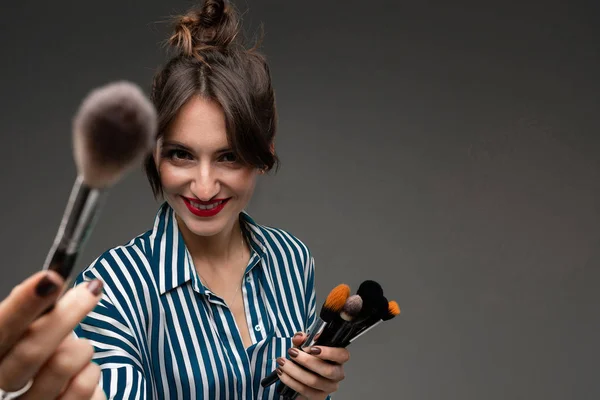 Portrait of young smiling woman in blue and white striped blouse with makeup brushes in her hands isolated. One brush is defocused. Photo with depth of field