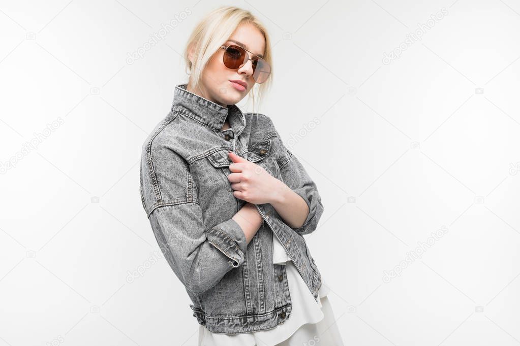 portrait of young charming joyful blonde girl in jeans jacket possing and gesticulating in round sunglasses on background
