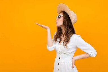 Stüdyoda beyaz elbise, hasır şapka ve güneş gözlüğü takmış, turuncuya karşı poz veren şık bir genç kadın.