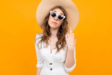 Stüdyoda beyaz elbise, hasır şapka ve güneş gözlüğü takmış, turuncuya karşı poz veren şık bir genç kadın.
