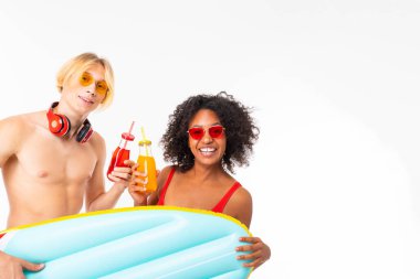African woman and Caucasian blonde man in swimwear posing with air mattress and beverages against white background 