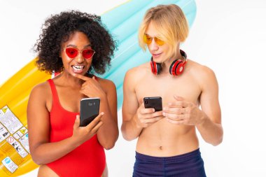 African woman and Caucasian blonde man in swimwear posing with mobile phones and air mattress against white background 
