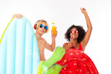 African woman and Caucasian blonde man in swimwear posing with air mattress and beverages against white background 