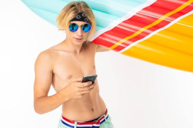 young blond man bare-chested in sunglasses posing with air mattress and mobile phone against white background  