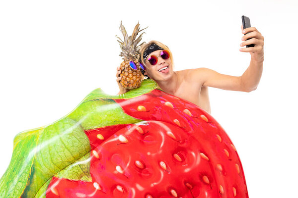 молодой блондин с голой грудью позирует с надувным матрасом и мобильным телефоном на белом фоне
  