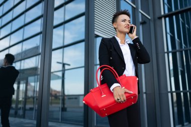 Modern binanın yakınında kırmızı çantalı resmi kıyafetler içinde poz veren güzel bir kadın.