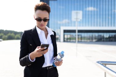 Güzel bir iş kadını telefon ve su şişesiyle modern binaya karşı poz veriyor.