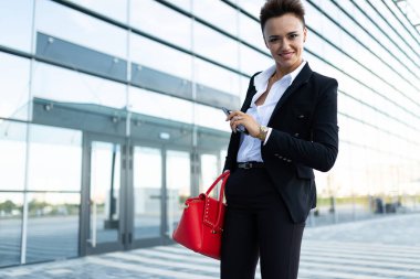 Modern binanın yakınında kırmızı çantalı resmi kıyafetler içinde poz veren güzel bir kadın.