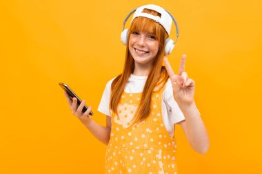 Kızıl saçlı, turuncu arka planda cep telefonu ve kulaklıkla poz veren güzel bir genç kız. 