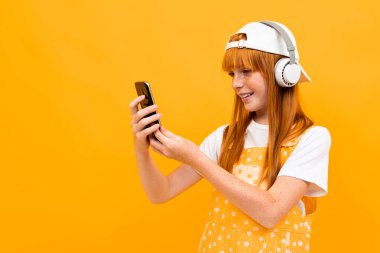 Kızıl saçlı, turuncu arka planda cep telefonu ve kulaklıkla poz veren güzel bir genç kız. 