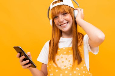 Kızıl saçlı, turuncu arka planda cep telefonu ve kulaklıkla poz veren güzel bir genç kız. 