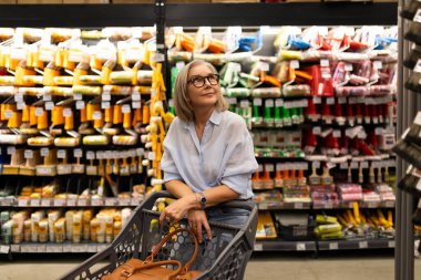 Gri saçlı ve gözlüklü bir kadın, bir marketin içindeki alışveriş arabasında oturuyor. Etrafı parlak renkli gıda ürünleri ve davetkar bir atmosferle çevrili..