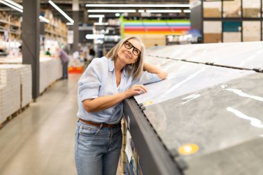 Gri saçlı ve gözlüklü bir kadın, bir mağazada çeşitli fayans seçimlerini inceliyor. Arka planda müşteriler arasında alışveriş yaparken düşünceli ve meşgul görünüyor..