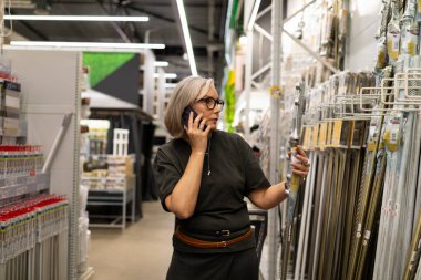 Bir kadın hırdavatçıda aletleri karıştırırken bir telefon görüşmesi yapar. Bir elindeki aleti inceliyor, ilgili ve odaklanmış görünüyor..