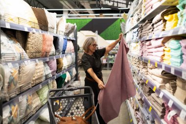 Bir müşteri iyi organize edilmiş bir ev eşyası mağazasında renkli yatak ve havlularla dolu koridorlarda gezinir ve ev dekorasyonu için özenle seçer..