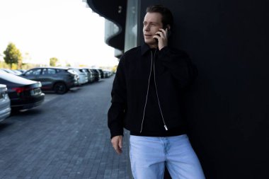 Genç bir adam zarif bir binaya karşı duruyor, telefon görüşmesi yapıyor. Güneşli bir günde etrafı park halindeki arabalarla çevrili siyah bir ceket ve açık renk kot giyiyor..