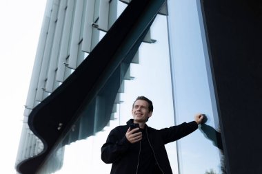 Bir adam parlak camdan bir binaya yaslanmış gülümsüyor ve akıllı telefonuna bakıyor. Parlak gün ışığı yapının çağdaş tasarımını vurgular.