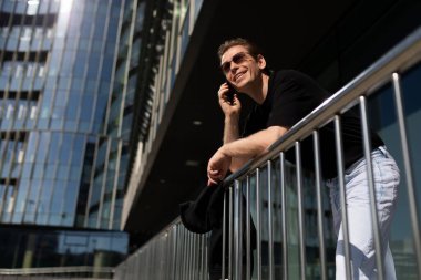 Bir adam güneşli bir günün tadını çıkarırken tırabzanlara yaslanır ve modern bir binanın önünde telefonla konuşur. Rahatlamış tavrı bir anlık bağ ve rahatlık yakalıyor..