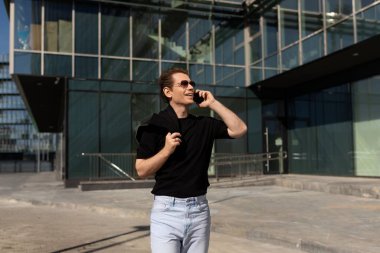 Bir adam güvenli bir şekilde yürür, açık gökyüzünün altında zarif, modern bir binanın dışında telefonla konuşur. Siyah gömlek ve açık renk kot pantolon giyiyor, sıradan ama şık bir hava yayıyor..