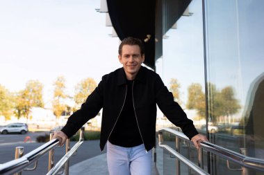 Çağdaş bir binanın dışındaki gümüş parmaklıklara yaslanmış bir adam gülümsüyor. Güneşli bir gün ve arka planda ağaçlar görülebiliyor, canlı atmosferi güçlendiriyor..