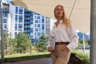 Beyaz gömlekli ve taba rengi pantolonlu bir kadın, elinde bir dizüstü bilgisayarla modern kentsel alanda geziniyor ve tepe örtülü bir yapının altında özenle ileriye bakıyor..