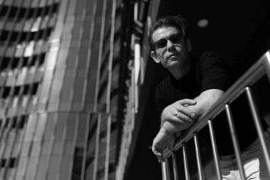 Güneş gözlüğü ve koyu renk gömlek takan bir adam korkulukların yanında gelişigüzel durur. Arka planda parlak güneş ışığını yansıtan, canlı bir şehir atmosferi yaratan yüksek ve modern binalar yer alıyor..