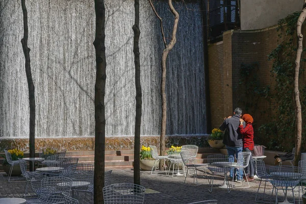 Paley Pocket Park 'taki şelaleyi izleyen erkek ve kadın, Manhattan şehir merkezinde, New York City' de, sarı lalelerin, beyaz masaların ve sandalyelerin arasında..