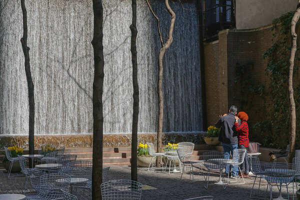 Man and woman watching the waterfall in Paley Pocket Park, midtown Manhattan, New York City, amid yellow tulips and white tables and chairs.