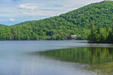 Adirondack Park, New York: Mavi Dağ Gölü 'nün kıyısından uzaktaki orman kaplı bir yamacın manzarası, bulutlar yukarıda, derin mavi bir gökyüzünde.