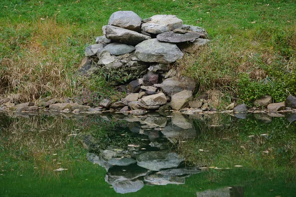 The mirror image of a pile of rocks on a grassy bank reflected in a stream.
