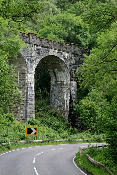 A beautiful Victorian-era stone arch bridge surrounded by a lush forest, at a curve in the road, in the Highlands of Scotland.