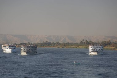 Nil Nehri, Mısır: Bir kayıkta iki adam Nil Nehri 'nin yukarısında akıntıya karşı giden birkaç nehir teknesini, arka planda ise batı yakasının kum tepelerini izliyor..