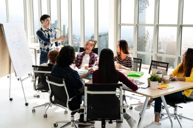 Genç Asyalı yaratıcı iş adamı modern ofiste durup sunum yapıyor. Takımla konuşup beyin fırtınası yapıyor. Sıradan insanlar iş görüşmesi konsepti.