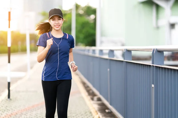 Young attractive asian runner woman running in city street or foot path way listen to music wearing blue or black sporty gadget in leisure sport concept with sun light in morning or evening workout.